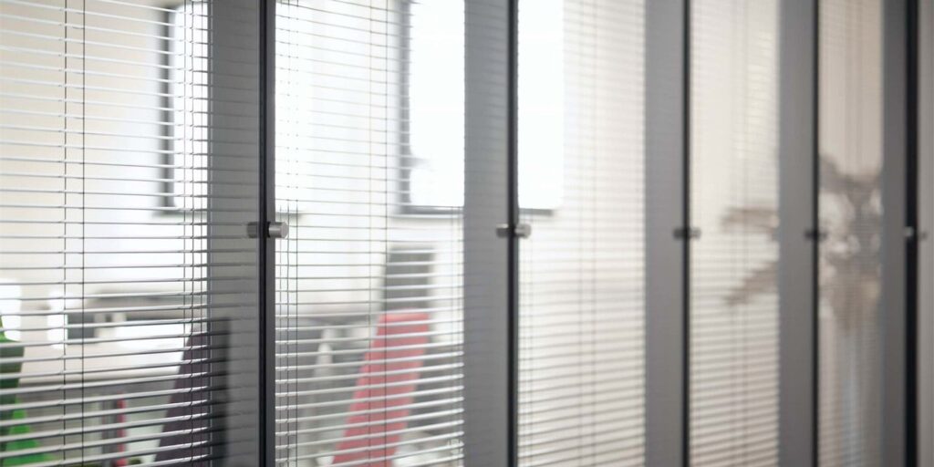 Grijze jaloezieën voor een kantoorraam met diffuus zicht op een rode bureaustoel.